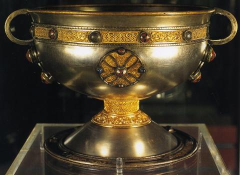 The Ardagh Chalice- a silver coloured large, wide chalice, with what appear to be rounded handles on either side, from the rim to just under half the height of the chalice's bowl (approx. quarter of the chalice's overall height. There are gold coloured intricate elements around the stem of the chalice, as a band encircling the bowl just beneath the rim, and as circles at 4 equidistant points around the bowl, with stones at the centre and at the top, bottom, left, and right of these circles